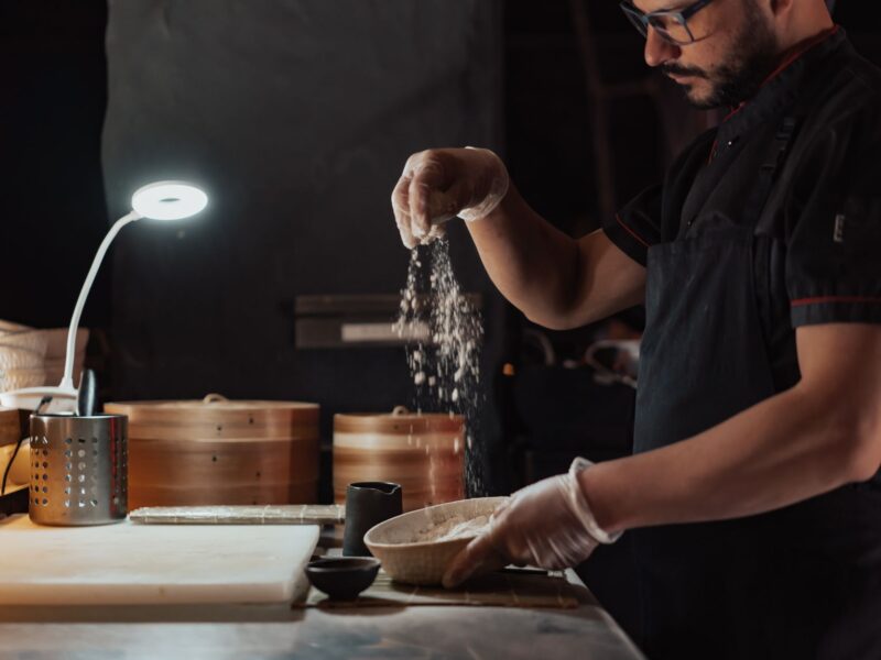 Chef wearing apron sprinkles flour over dish in a dimly lit kitchen setting, emphasizing traditional cooking.