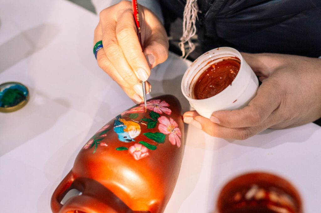 Close-up of an artist painting a colorful bird on a clay vase, showcasing intricate detail.